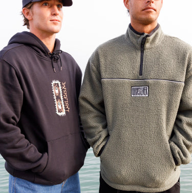 Two men in a black hoodie and polar fleece quarter zip stand together by the ocean, with waves in the background.