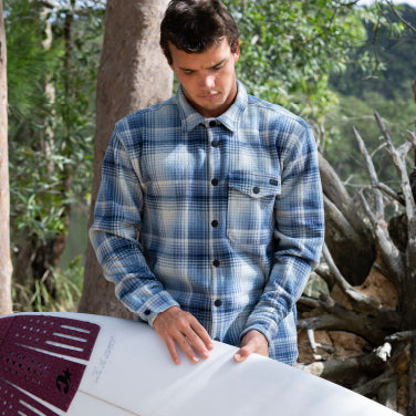 A man in a plaid flannel shirt holds a surfboard, ready for a day at the beach